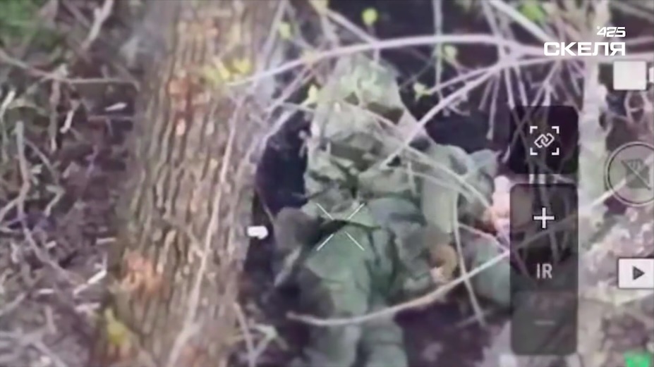 Russian lies belly up on the ground in a tree line right after receiving FPV hit. He takes his ballistic vest off, grabs a grenade, installs or changes fuse on it, and detonates it near his chest