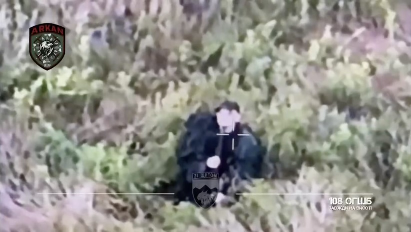 Russian receives multiple drone drops while moving across open field, later is seen sitting in high grass with baseball cap on. He fumbles with his rifle, which appears to be jammed. Finally, after several test shots he shoots himself sending his cap off